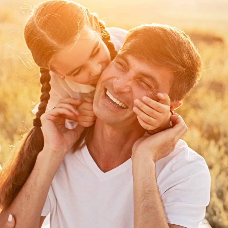 Daughter hugging and kissing cheerful father in cheek near laughing mother while spending day of happy childhood in field